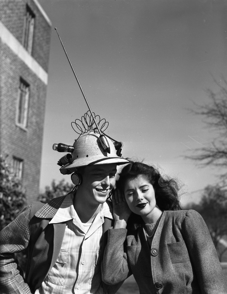 A photo of Olin Mumford wearing his radio hat. A woman listens in.
